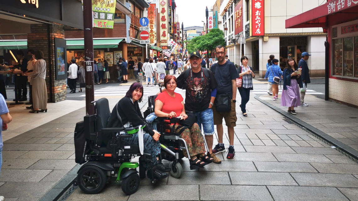 Four people, two in wheelchairs, pose on a busy street in Tokyo lined with shops and restaurants. Pedestrians and colorful signs fill the background.