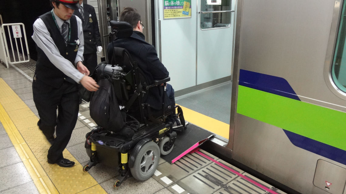 A man in a power wheelchair is going up a portable ramp placed by a staff member who helps push him into a subway