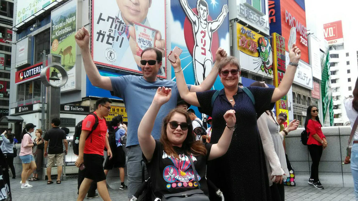 A cheerful group of three posing in front of the Glico Running Man sign in Dotonbori, Osaka. The background is filled with colorful billboards and people.