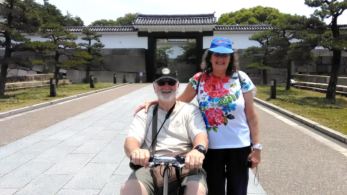 An older couple smiling in front of Osaka Castle. The man is in a wheelchair, and both wear hats. Pine trees and castle walls are in the background.