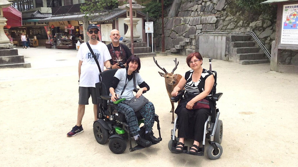 A group of four, two in wheelchairs, pose with a friendly deer in Miyajima, Japan, near shops and stone steps.