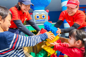 Children and staff build with large LEGO bricks at Legoland Japan, smiling and having fun near a LEGO robot figure.