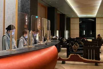 Three masked staff work behind a curved hotel reception desk with clear partitions in a dimly lit, modern lounge with wooden decor.