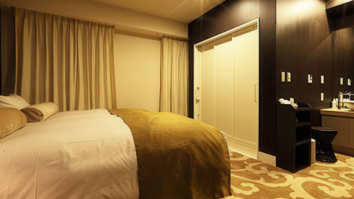 A cozy hotel room with a large bed covered in white and gold bedding, thick curtains, and a small vanity area near a closet.