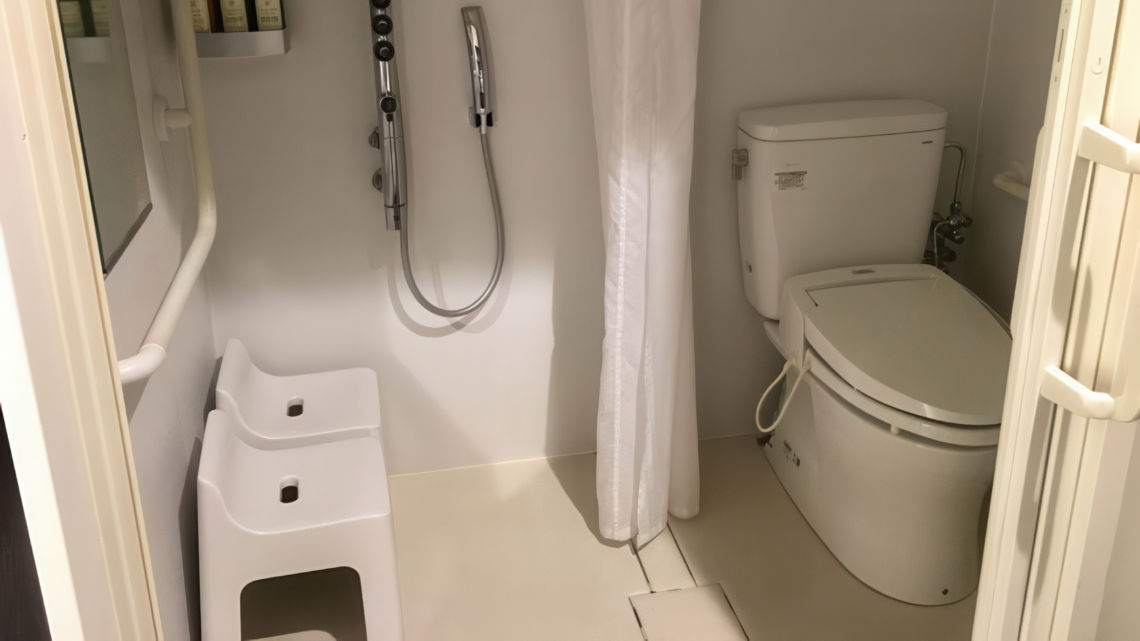 A compact bathroom with a toilet, handheld shower, and two white stools under a showerhead, all in a minimalistic, white design.