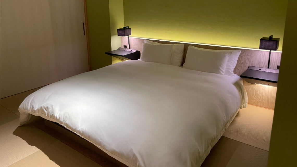 A minimalist hotel room with a large, neatly made bed, flanked by two wall-mounted nightstands with lamps, and a soft green accent wall.