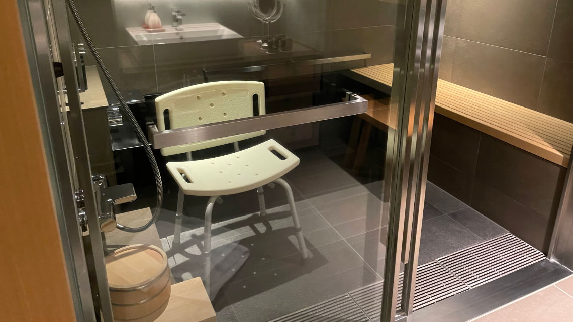 A modern bathroom with a shower area featuring a white shower chair, wooden bench, and a bucket, all behind glass doors.