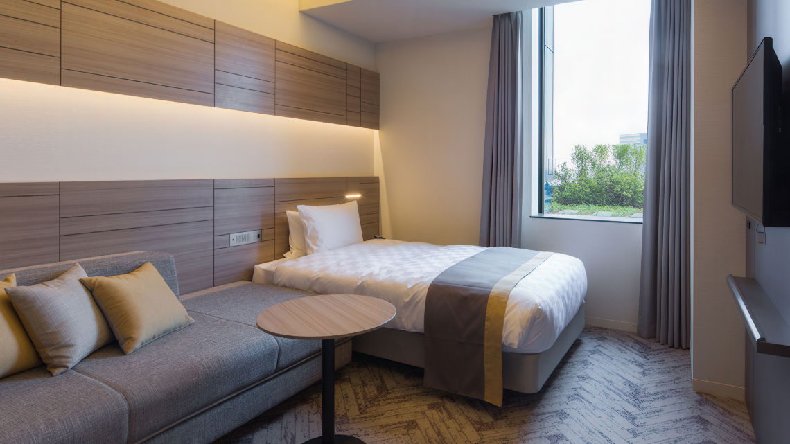 A bright hotel room with a single bed, gray sofa, small round table, and a large window with curtains, featuring wooden accents and soft lighting.