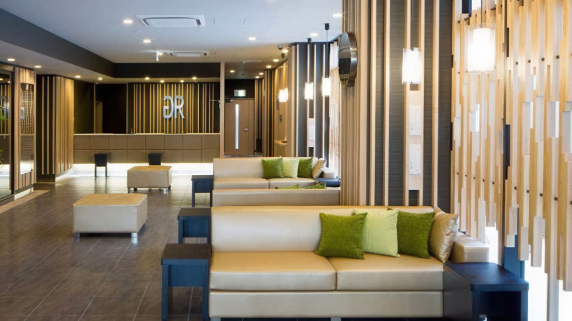 A stylish hotel lobby with beige sofas, green accent pillows, wooden paneling, and soft lighting, leading to a modern reception area.