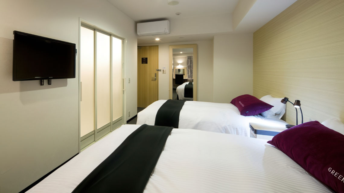 A compact, modern hotel room with two single beds, purple pillows, a wall-mounted TV, and frosted sliding doors near the entrance.