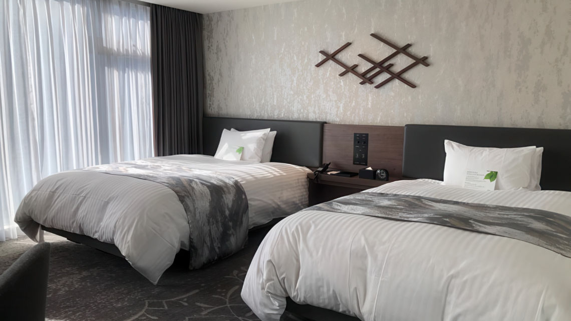 A contemporary hotel room with two neatly made beds, abstract wall art, soft lighting, and floor-to-ceiling curtains.