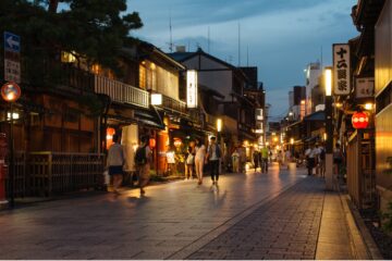 A lively, lantern-lit street in Japan at dusk, with traditional wooden buildings and pedestrians strolling along the cobblestone path.