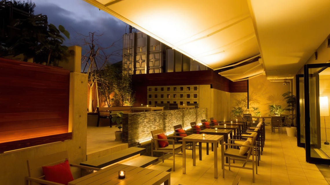 A modern outdoor terrace at night with neatly arranged tables, warm lighting, and a cozy ambiance, featuring candles and red cushions.