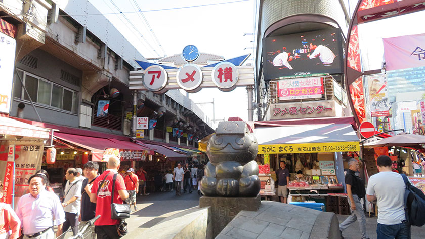 A busy outdoor shopping street in Japan with the "Ameyoko" sign, people walking, shops, and a stone statue in the center.