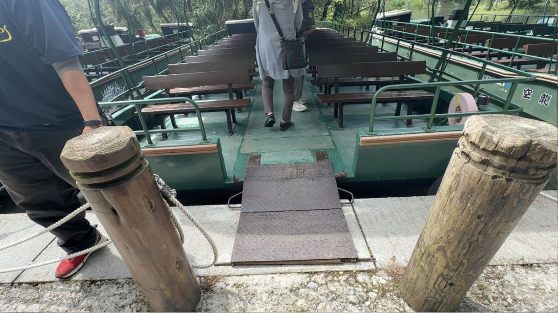 Two people boarding a boat with wooden benches via a small ramp, with dock posts and ropes in the foreground.