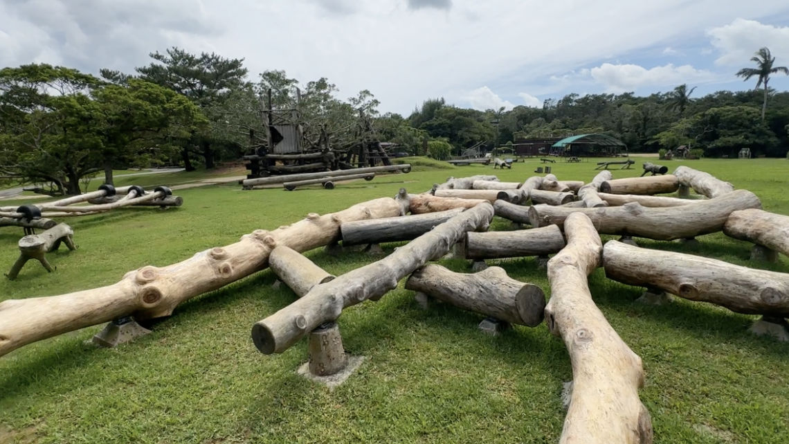 Outdoor playground with large wooden logs arranged on grass, surrounded by trees and picnic benches in the distance.