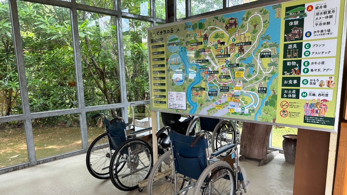 Three wheelchairs are parked in front of a colorful map inside a glass-walled area with greenery outside.