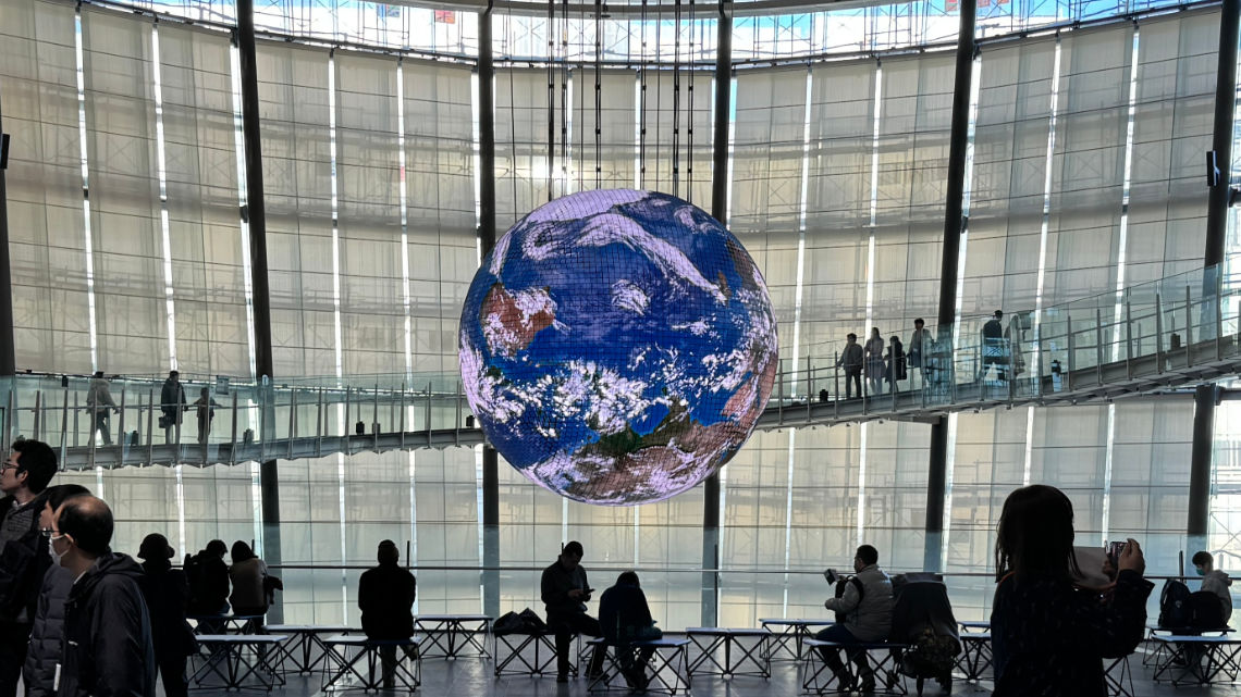 People observe a large digital globe display suspended in a modern indoor space with a glass walkway and high ceilings.