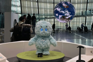 A fluffy blue mascot stands on display in front of an audience, with a large digital globe hanging in the background of a modern exhibit space.