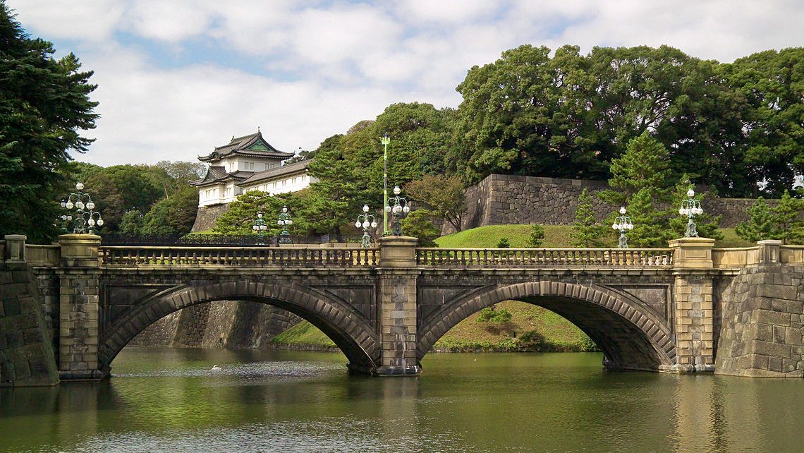 A stone bridge spans a moat in front of Japan's Imperial Palace, surrounded by lush greenery and traditional architecture.