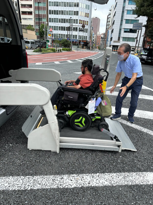 Tayjus Surampudi in his power wheelchair being assisted onto a wheelchair-accessible van via a lift in Tokyo. A staff member ensures safety as they prepare for transport through the city.