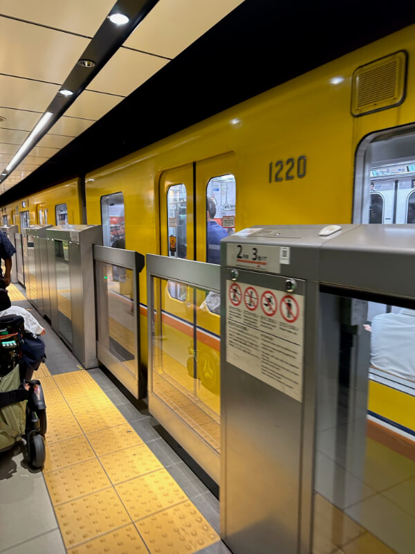 Tayjus Surampudi waits near the platform in his power wheelchair beside a yellow Tokyo Metro train. The platform is equipped with automatic safety barriers, ensuring a secure boarding experience.