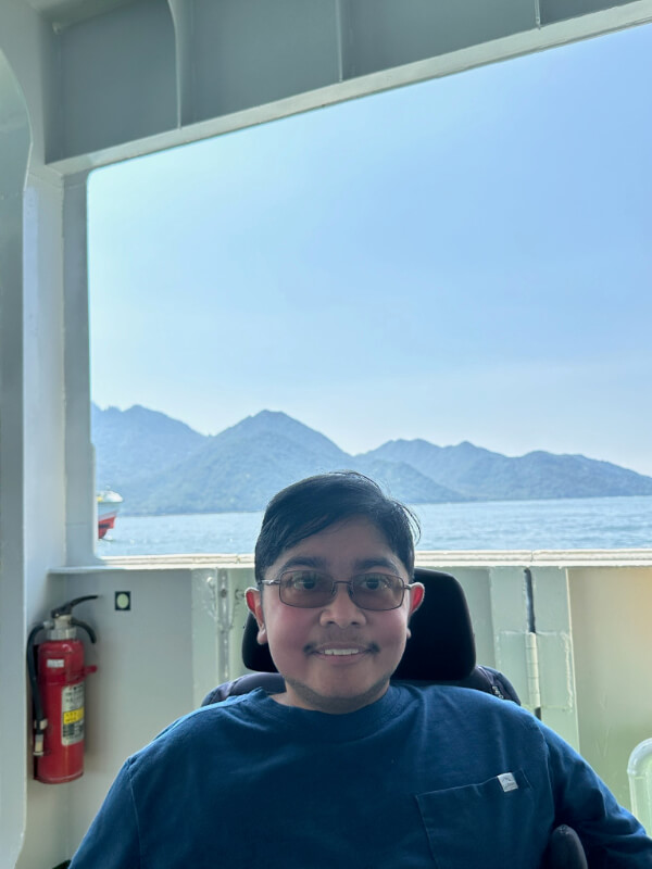 Tayjus Surampudi enjoys the scenic view of Miyajima Island from the ferry. The mountains and calm waters are visible in the background as he travels in his power wheelchair.