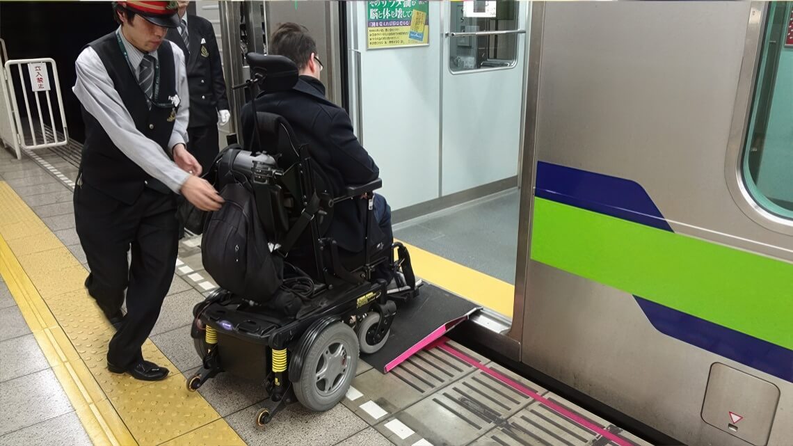 A train station staff member assists a man in a motorized wheelchair onto a train using a portable ramp at the platform.