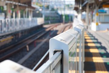 An empty train station platform with safety gates closed along the tracks. No train is visible, and sunlight illuminates the scene.