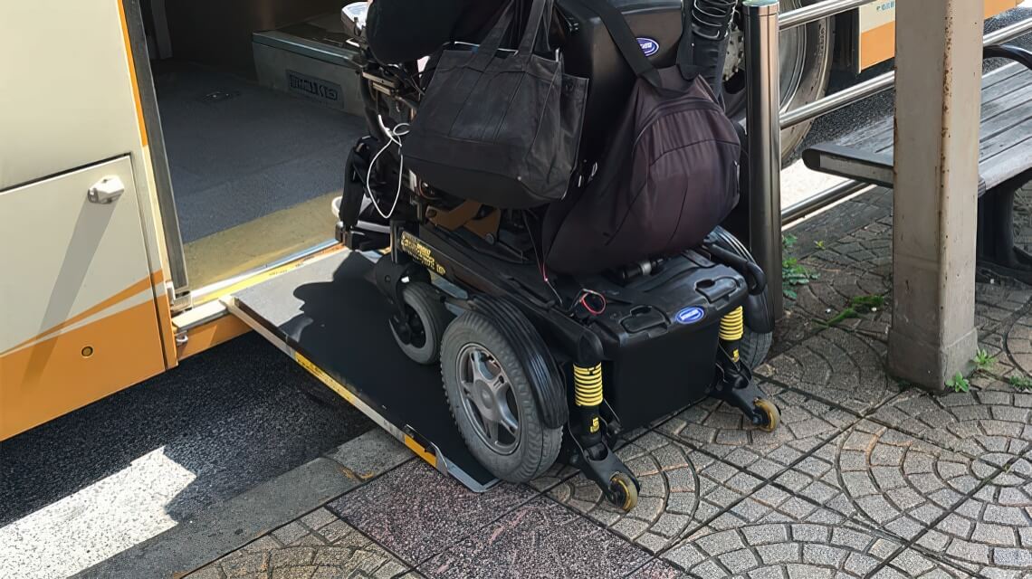 A person in a motorized wheelchair boards a bus using a deployed ramp at the bus stop. The wheelchair carries bags on the back.