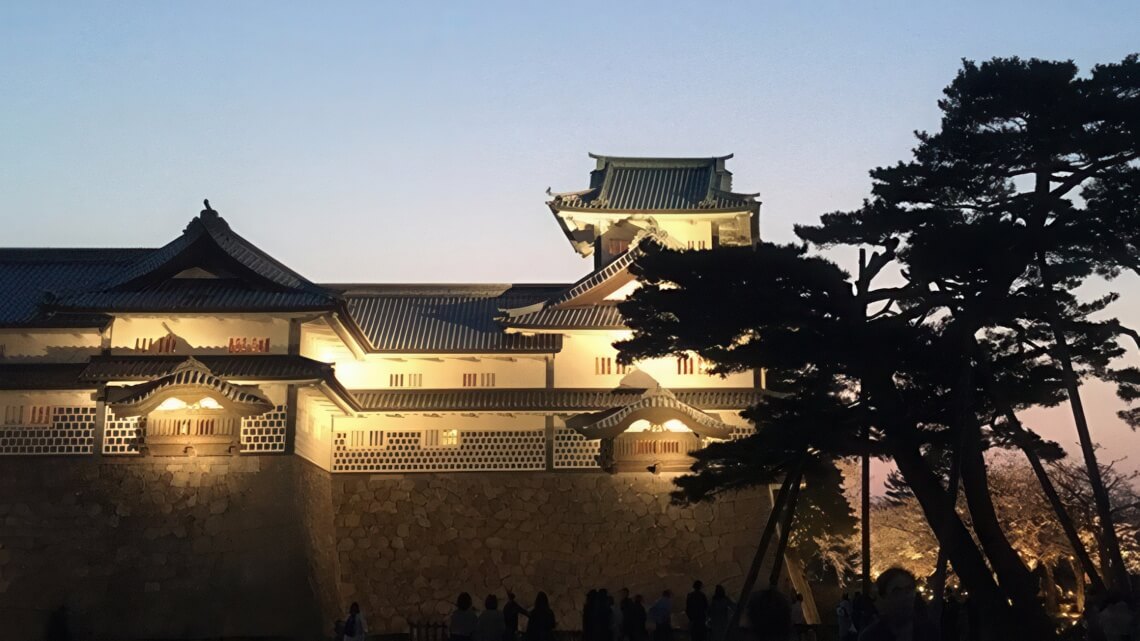 Kanazawa Castle illuminated at dusk, showcasing its traditional architecture with silhouetted pine trees in the foreground.