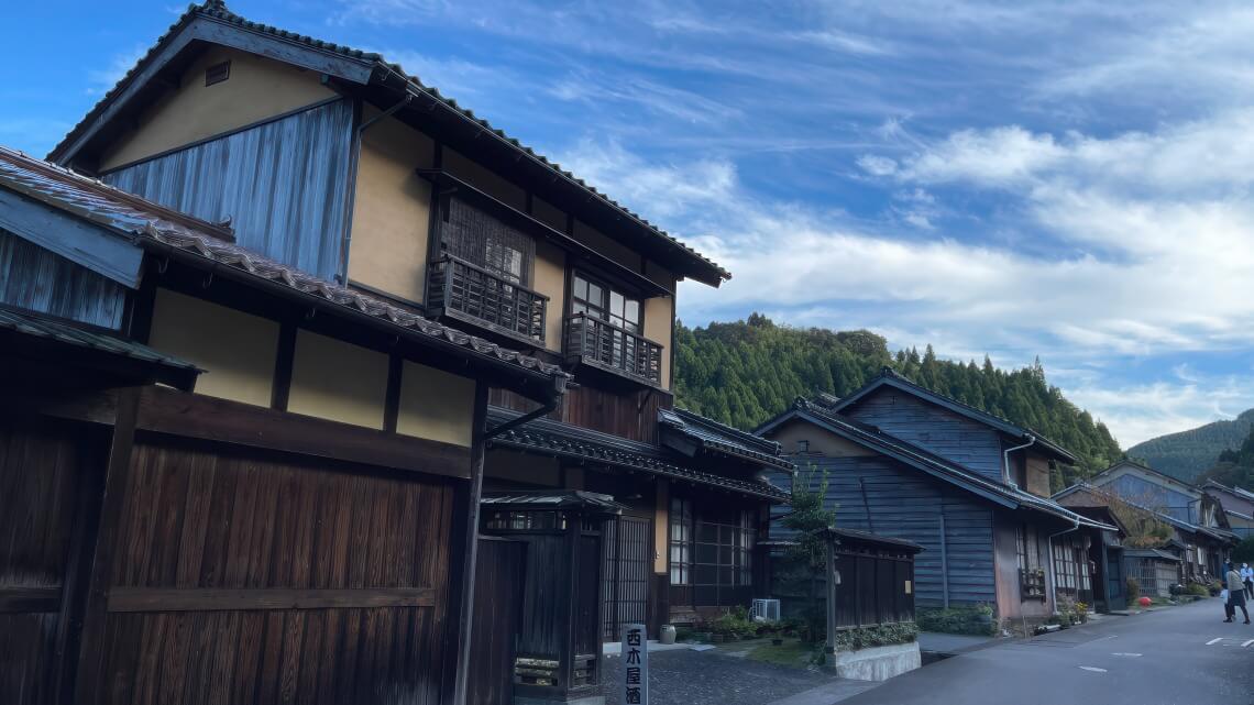 Traditional wooden houses line a quiet street in a rural Japanese village, with forested hills and a blue sky in the background.