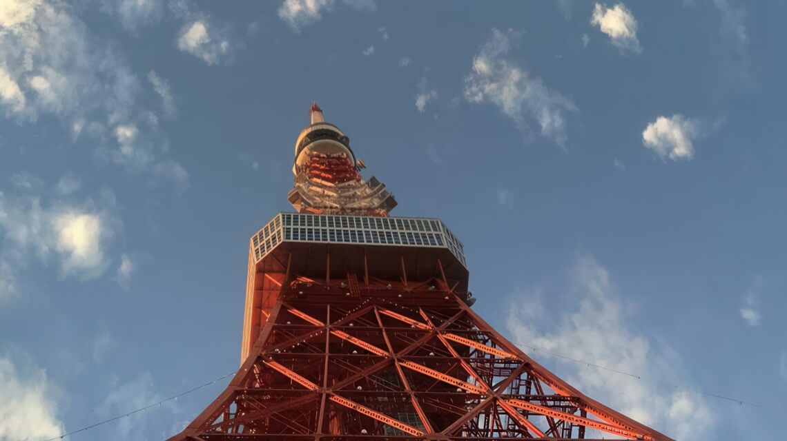 Upward view of Tokyo Tower's red steel structure and observation deck against a blue sky with scattered clouds.