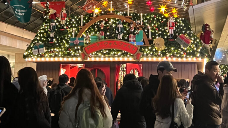 A festive Christmas market stall decorated with nutcrackers, gifts, candles, and lights, surrounded by people in warm winter clothing.