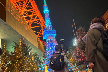 Two people admire Tokyo Tower at night, lit in bright blue and green, surrounded by decorated Christmas trees with twinkling lights.