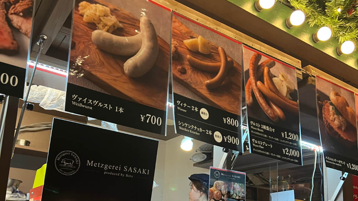 A food stall menu displaying various sausages, including Weißwurst and Wiener, with prices ranging from ¥700 to ¥2,000, under festive lights.