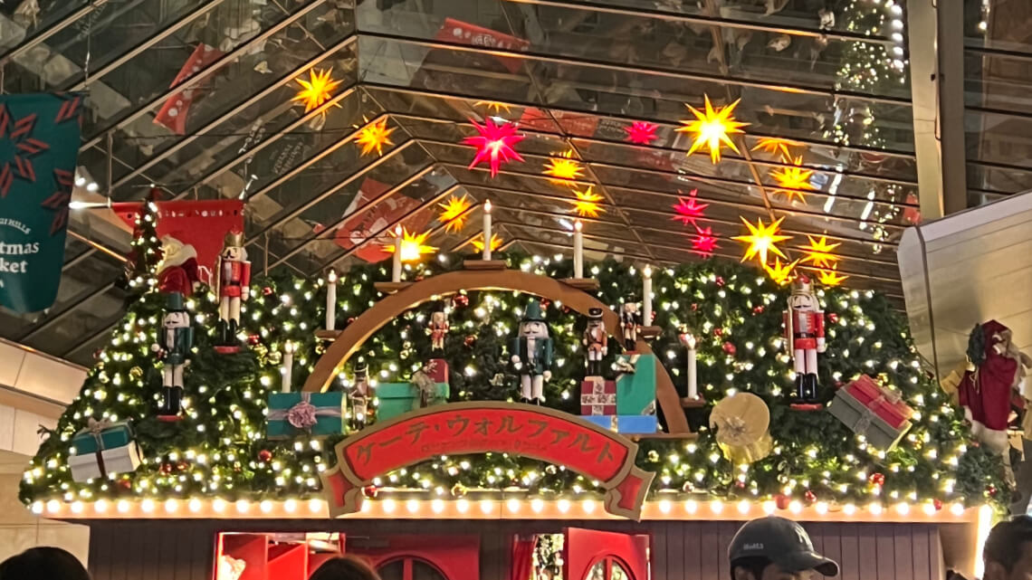 A festive Christmas market stall adorned with nutcrackers, candles, wrapped gifts, and string lights under a glass roof with star lights.