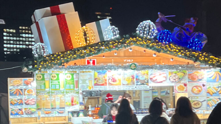 A festive food stall decorated with oversized gift boxes, lights, and garlands, serving colorful drinks and snacks at a holiday market.