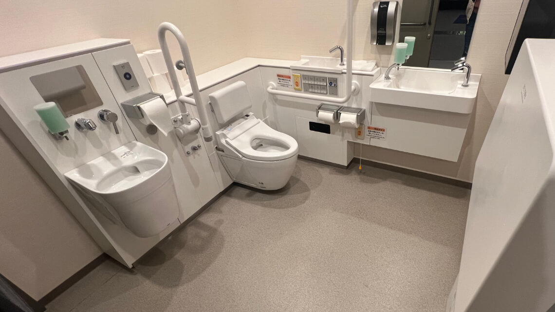 A spacious and modern accessible restroom with a bidet toilet, safety handrails, a support seat, and an emergency pull cord. The room includes two sinks—one at a lower height for wheelchair users the other for ostomate users.