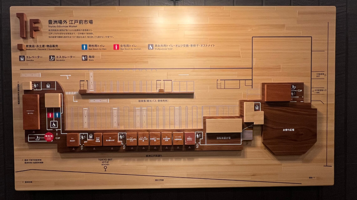 A detailed wooden 3D map of Toyosu Edo-Mae Market's first floor, showing stores, facilities, restrooms, and access points. Clear icons indicate elevators, escalators, male, female, and multi-purpose toilets. Labels in Japanese and English.