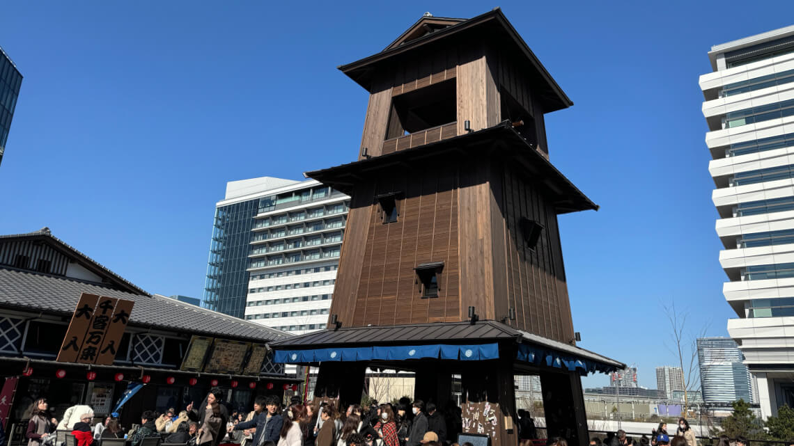 A tall wooden bell tower at Toyosu Senkyaku Banrai stands prominently under a bright blue sky. Crowds gather around its base, enjoying the lively atmosphere. Traditional-style shopfronts with signs and blue awnings surround the area, while modern high-rise buildings rise in the background.