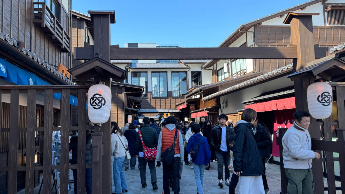 A bustling pathway at Toyosu Senkyaku Banrai lined with traditional wooden buildings featuring tiled roofs and colorful awnings. A wooden arch with lanterns bearing Japanese patterns frames the entrance.