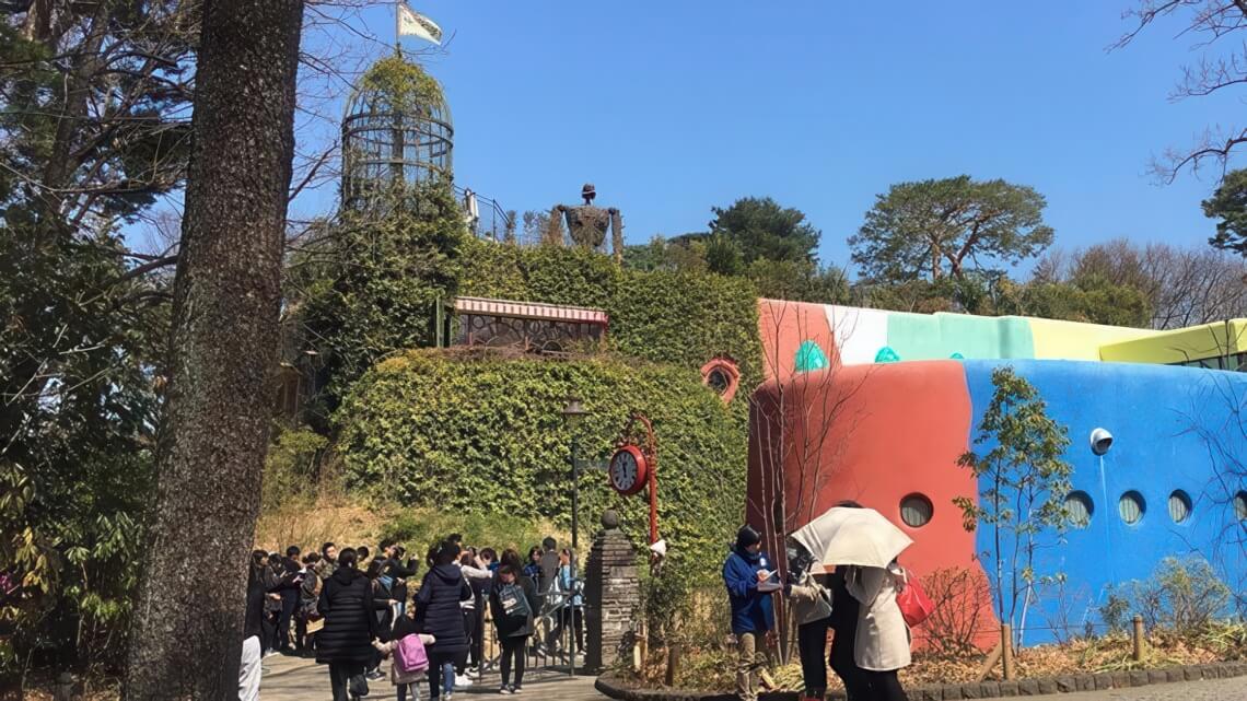 Visitors gather outside of the Ghibili Museum, a colorful building with curved walls and a rooftop garden, featuring a large robot statue in the background.