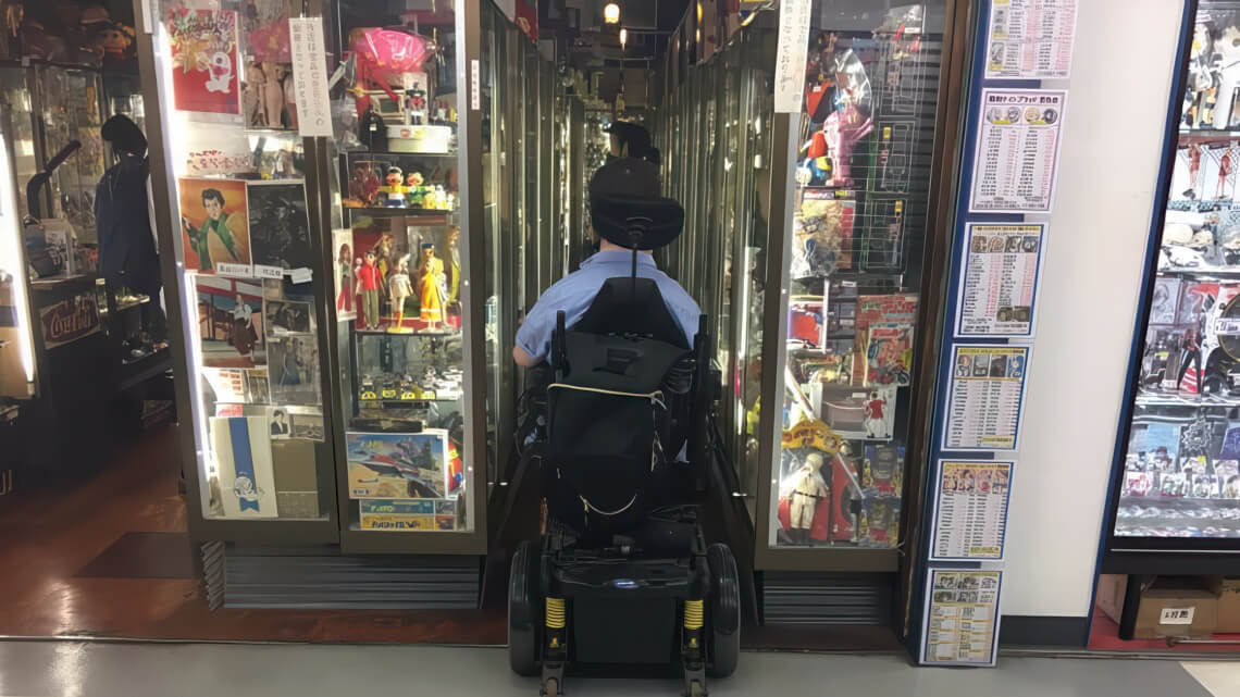 A person in a motorized wheelchair navigates a narrow aisle lined with display cases filled with toys and collectibles.