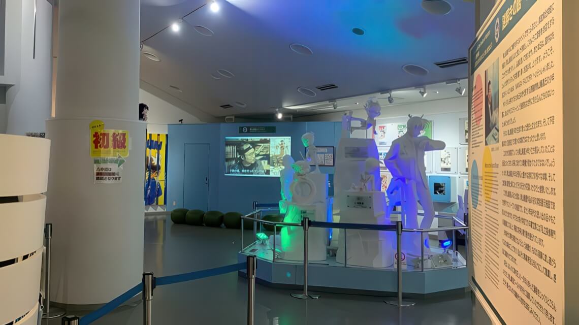 A museum exhibit with illuminated white statues, informational panels, and a video projection on a blue wall in the background.