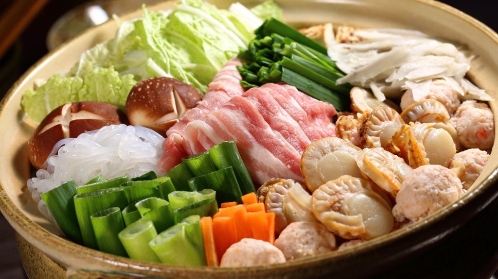 A beautifully arranged Japanese hot pot (nabe) featuring fresh ingredients such as napa cabbage, shiitake mushrooms with cross-cut patterns, sliced pork, scallops, fish balls, glass noodles, green onions, and julienned carrots. The vibrant vegetables and proteins are presented in a circular pattern in a clay pot, ready for cooking.