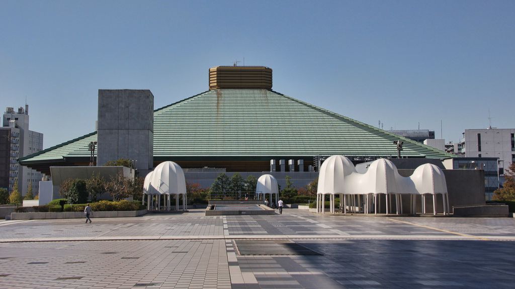 Ryogoku Kokugikan, a large sumo wrestling arena in Tokyo, Japan, with a distinctive green pyramid-shaped roof and a golden top structure. The building is surrounded by a wide open plaza featuring geometric tile patterns, small trees, and modern white structures resembling sculptures or canopies. The clear blue sky enhances the architecture's prominence.