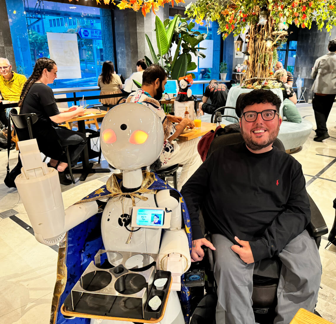 Cory Lee, a wheelchair user, seated next to a robot server at DAWN Avatar Robot Cafe in Tokyo, with other patrons dining in the background