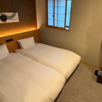 The secondary bedroom of the Two Bedroom Theater Suite has two twin beds with an art piece on the wall, Japanese style-window covering, stylish lamp, flatscreen TV on the wall and a closet.