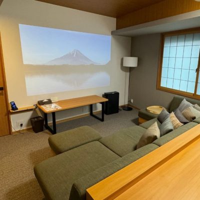 The Two Bedroom Theater Suite Living area has a large couch with throw pillows and ottoman footrests. There Japanese-style paper blinds on the windows and a projector for watching TV or movies on the wall.